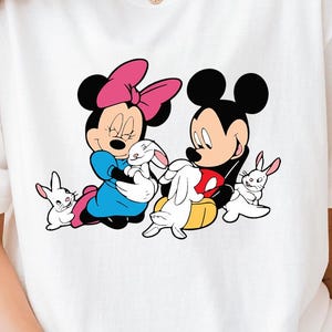 May include: White t-shirt featuring a cartoon image of Mickey Mouse and Minnie Mouse holding a white bunny rabbit. The characters are surrounded by three smaller white bunny rabbits.