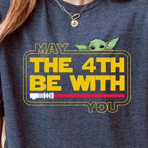 May include: A heather navy blue t-shirt with a graphic of a baby Yoda peeking out from behind a yellow banner that reads "May the 4th be with you". The shirt has a "Comfy" logo on the bottom.