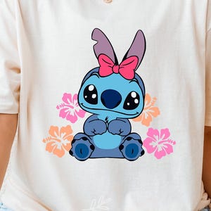 May include: A white t-shirt with a blue Stitch character wearing a pink bow and surrounded by pink and yellow flowers. The text "Natural" is visible in the bottom right corner of the image.
