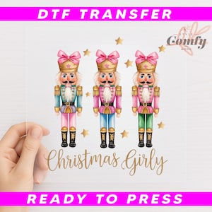 May include: A clear DTF transfer sheet featuring three colorful nutcracker soldier illustrations. Each soldier has a pink bow, gold hat, and a different colored uniform: blue, pink, and green. The text "Christmas Girly" is written in gold script.