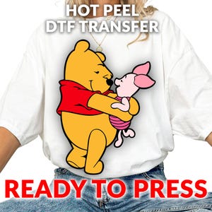 May include: White t-shirt featuring a cartoon image of Winnie the Pooh hugging Piglet. Winnie the Pooh is yellow and wearing a red shirt. Piglet is pink and wearing a pink and white striped shirt. Text at the top reads "HOT PEEL DTF TRANSFER" and at the bottom "READY TO PRESS".