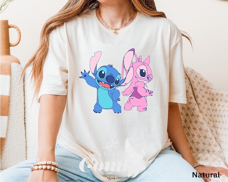 May include: Cream-colored t-shirt featuring a blue cartoon character waving and a pink cartoon character. The shirt has the word "Natural" printed on the bottom right corner. The shirt is a short-sleeved style.