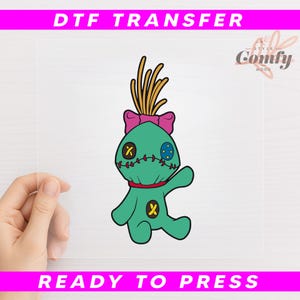 May include: A DTF transfer featuring a cartoon doll with a teal body, a pink bow, and yellow hair. The doll has stitched details and a red neck. The text "DTF TRANSFER" and "READY TO PRESS" are displayed in pink.