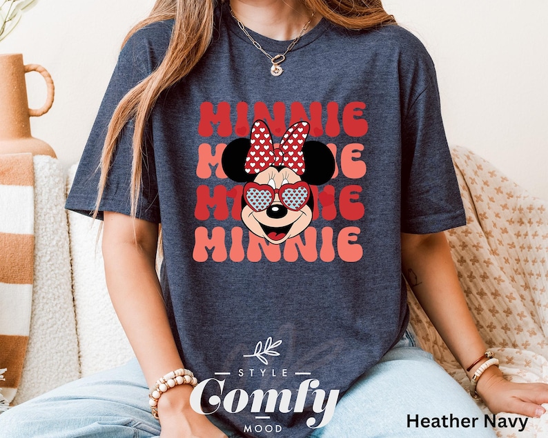 Puede incluir: Una camiseta azul marino jaspeada con un gr&aacute;fico de Minnie Mouse con gafas de sol en forma de coraz&oacute;n y un lazo rojo con lunares blancos. El texto "Minnie" se repite en una fuente retro en tonos de rosa y rojo.