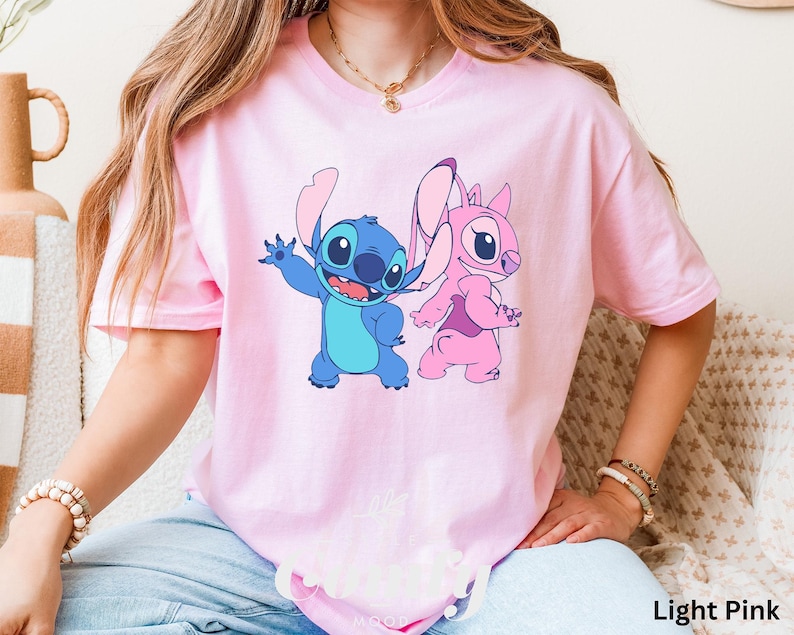 May include: Light pink t-shirt featuring a cartoon graphic of Stitch and Angel. Stitch is blue, and Angel is pink. The text "Light Pink" is at the bottom right of the image. The shirt has short sleeves and a crew neck.