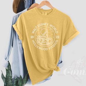 Puede incluir: Una camiseta amarilla con un gr&aacute;fico blanco de un c&iacute;rculo con el texto "Walt Disney World Hollywood Studios 20 22" y una imagen estilizada de un castillo.