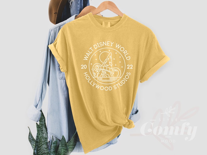 Puede incluir: Una camiseta amarilla con un gr&aacute;fico blanco de un c&iacute;rculo con el texto "Walt Disney World Hollywood Studios 20 22" y una imagen estilizada de un castillo.