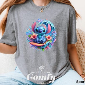 Może przedstawiać: Szara koszulka sportowa z kolorową grafiką postaci Stitch z filmu Lilo & Stitch. Stitch jest przedstawiony na ż&oacute;łtej desce surfingowej, otoczony falą i kwiatami. Koszulka ma napis "Style Comfy Mood".