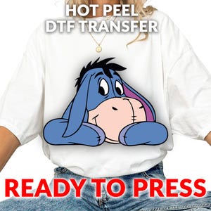 May include: White t-shirt featuring a blue Eeyore graphic. The image includes the text "HOT PEEL DTF TRANSFER" at the top and "READY TO PRESS" at the bottom. The Eeyore graphic is in the center of the shirt.