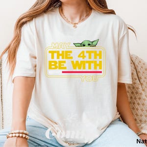 May include: A white t-shirt with a yellow graphic that says "May the 4th Be With You" and features a green alien character peeking out from behind the text.