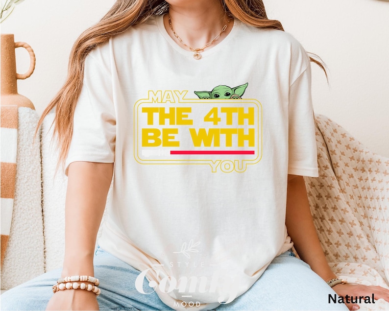 May include: A white t-shirt with a yellow graphic that says "May the 4th Be With You" and features a green alien character peeking out from behind the text.