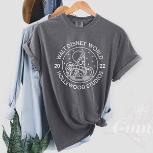 Puede incluir: Una camiseta gris oscuro con un dise&ntilde;o gr&aacute;fico blanco en forma de c&iacute;rculo con el texto "Walt Disney World 20 Hollywood Studios 22" y una ilustraci&oacute;n estilizada del Hollywood Tower Hotel.