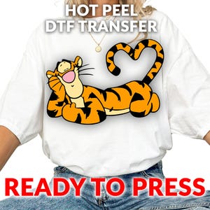 May include: White t-shirt featuring a cartoon tiger character in orange and black stripes, with a heart-shaped tail. The text "HOT PEEL DTF TRANSFER" and "READY TO PRESS" are printed on the shirt.