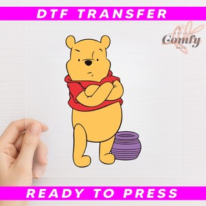 May include: A DTF (Direct to Film) transfer featuring a cartoon illustration of Winnie the Pooh with arms crossed, wearing a red shirt. A purple pot is next to him. The text "DTF TRANSFER" and "READY TO PRESS" are displayed.