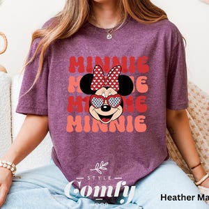 Puede incluir: Una camiseta de color marr&oacute;n rojizo con un gr&aacute;fico retro de Minnie Mouse con gafas de sol en forma de coraz&oacute;n y un lazo rojo. El texto "Minnie" se repite tres veces en una fuente retro en capas.