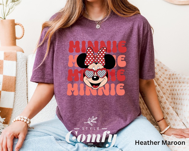 Puede incluir: Una camiseta de color marr&oacute;n rojizo con un gr&aacute;fico retro de Minnie Mouse con gafas de sol en forma de coraz&oacute;n y un lazo rojo. El texto "Minnie" se repite tres veces en una fuente retro en capas.