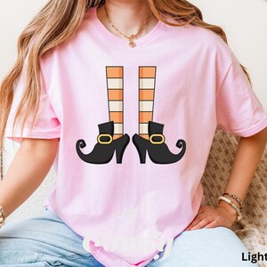 May include: Light pink t-shirt featuring a graphic of witch legs. The design includes black shoes with gold buckles and orange and white striped stockings. The text "Light Pink" is at the bottom right of the image.