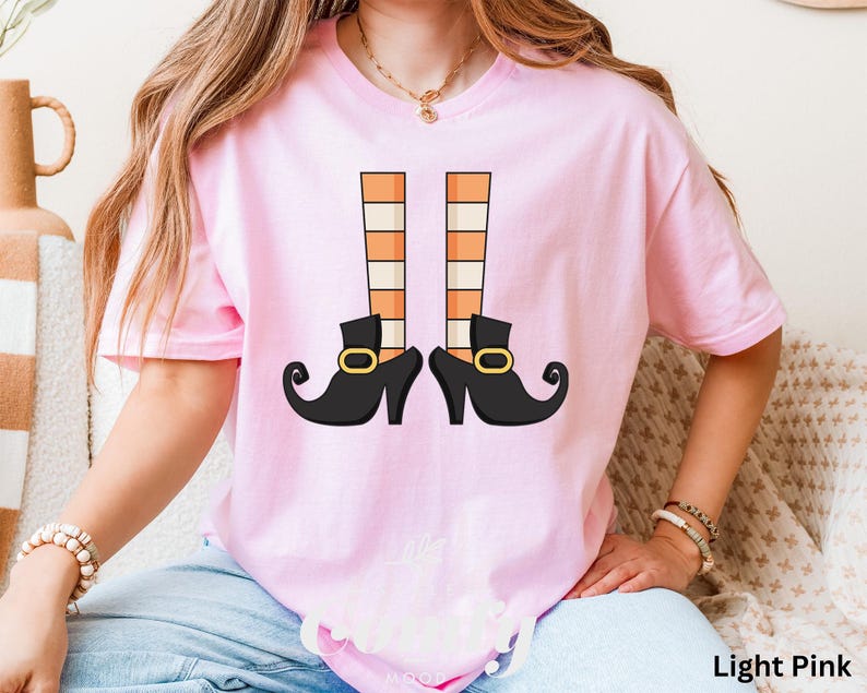 May include: Light pink t-shirt featuring a graphic of witch legs. The design includes black shoes with gold buckles and orange and white striped stockings. The text "Light Pink" is at the bottom right of the image.