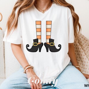 May include: White t-shirt featuring a whimsical illustration of witch legs. The design includes black shoes with gold buckles and orange and beige striped stockings. The word "Comfy" is printed on the shirt.