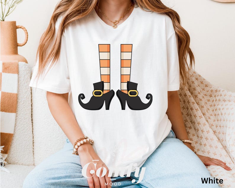 May include: White t-shirt featuring a whimsical illustration of witch legs. The design includes black shoes with gold buckles and orange and beige striped stockings. The word "Comfy" is printed on the shirt.