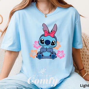 May include: Light blue t-shirt with a cartoon Stitch character wearing a pink bow and surrounded by pink and orange flowers. The shirt has the text "Comfy" printed on the bottom.