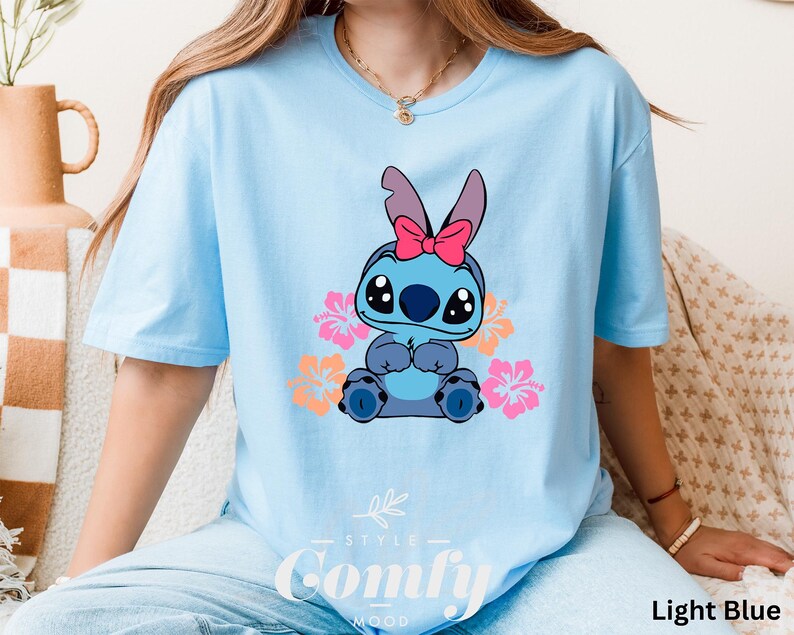 May include: Light blue t-shirt with a cartoon Stitch character wearing a pink bow and surrounded by pink and orange flowers. The shirt has the text "Comfy" printed on the bottom.