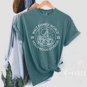 Puede incluir: Camiseta azul turquesa con un dise&ntilde;o gr&aacute;fico blanco en forma de c&iacute;rculo con el texto "Walt Disney World 20 Hollywood Studios 22" y una imagen estilizada del icono de Hollywood Studios.