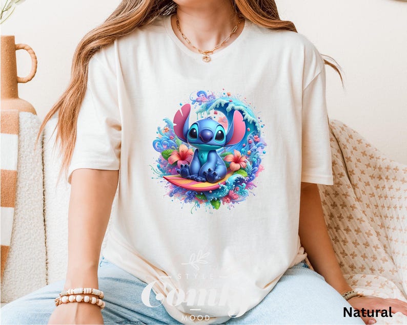 Może przedstawiać: Kremowy t-shirt z kolorową grafiką postaci Stitch z Lilo & Stitch. Stitch jest przedstawiony surfujący na desce surfingowej, otoczony falą i kwiatami. Słowo "Natural" jest wydrukowane w prawym dolnym rogu.