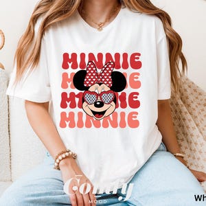 Puede incluir: Camiseta blanca con un gr&aacute;fico de Minnie Mouse rojo y blanco. El gr&aacute;fico presenta a Minnie Mouse usando gafas de sol en forma de coraz&oacute;n y un lazo rojo con lunares blancos. El texto "MINNIE" se repite en una fuente retro en rojo y rosa.