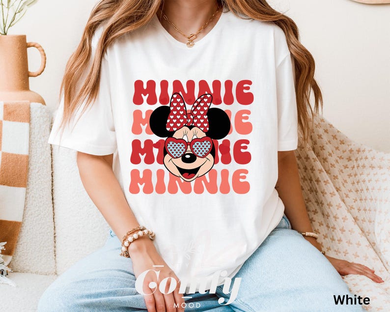 Puede incluir: Camiseta blanca con un gr&aacute;fico de Minnie Mouse rojo y blanco. El gr&aacute;fico presenta a Minnie Mouse usando gafas de sol en forma de coraz&oacute;n y un lazo rojo con lunares blancos. El texto "MINNIE" se repite en una fuente retro en rojo y rosa.
