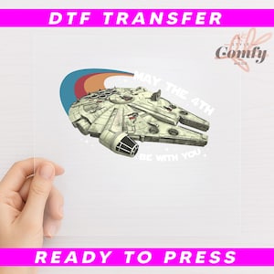 May include: DTF transfer featuring a detailed illustration of the Millennium Falcon starship, with the text "MAY THE 4TH BE WITH YOU." The design includes a retro-style arc with red, orange, and blue colors. The words "DTF TRANSFER" and "READY TO PRESS" are also visible.