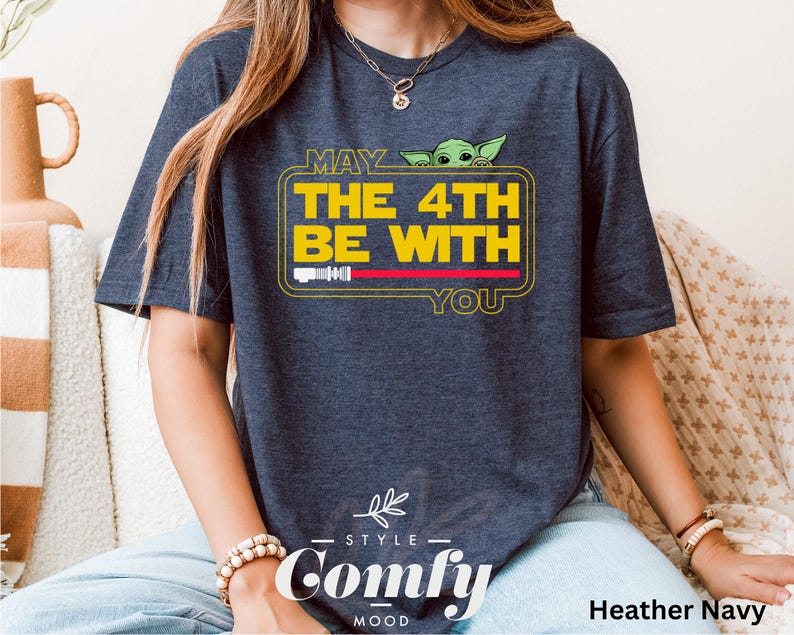 May include: A heather navy blue t-shirt with a graphic of a baby Yoda peeking out from behind a yellow banner that reads "May the 4th be with you". The shirt has a "Comfy" logo on the bottom.
