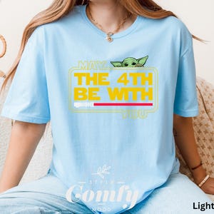 May include: Light blue t-shirt with a graphic of a green alien character peeking out from behind a yellow box with the text "May the 4th Be With You".