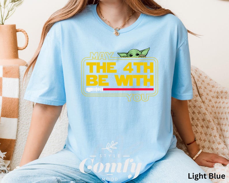 May include: Light blue t-shirt with a graphic of a green alien character peeking out from behind a yellow box with the text "May the 4th Be With You".