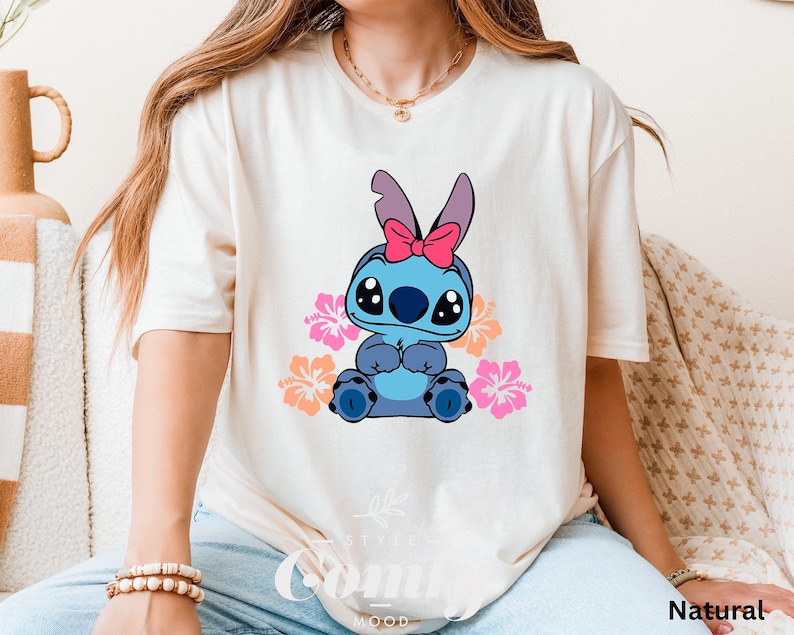 May include: A white t-shirt with a blue Stitch character wearing a pink bow and surrounded by pink and yellow flowers. The text "Natural" is visible in the bottom right corner of the image.