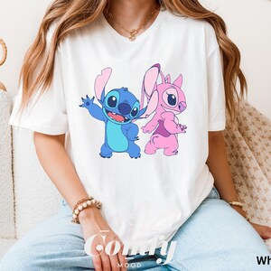 May include: White t-shirt featuring a cartoon graphic of Stitch and Angel. Stitch is blue with large eyes and Angel is pink. The word "Comfy" is printed in cursive on the bottom left of the shirt, and the word "White" is printed on the bottom right.