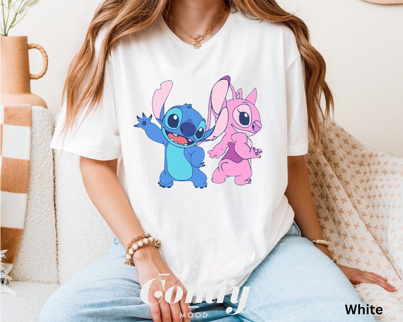 May include: White t-shirt featuring a cartoon graphic of Stitch and Angel. Stitch is blue with large eyes and Angel is pink. The word "Comfy" is printed in cursive on the bottom left of the shirt, and the word "White" is printed on the bottom right.