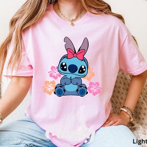 May include: A light pink t-shirt with a blue Stitch character wearing a pink bow. Stitch is surrounded by pink, orange, and yellow flowers. The text "Light Pink" is visible in the bottom right corner of the image.