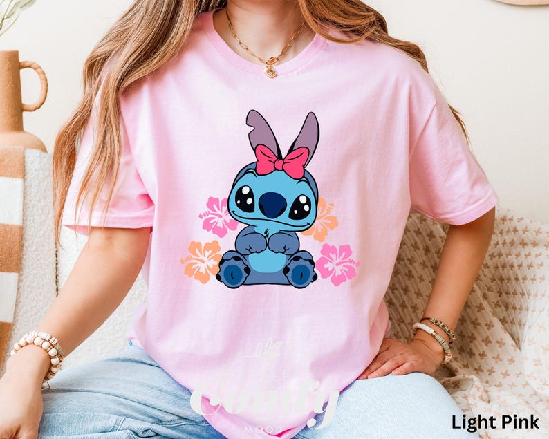 May include: A light pink t-shirt with a blue Stitch character wearing a pink bow. Stitch is surrounded by pink, orange, and yellow flowers. The text "Light Pink" is visible in the bottom right corner of the image.