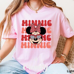 Puede incluir: Una camiseta rosa claro con un gr&aacute;fico retro de Minnie Mouse con gafas de sol en forma de coraz&oacute;n y un lazo rojo. El texto "MINNIE" se repite en un patr&oacute;n apilado.