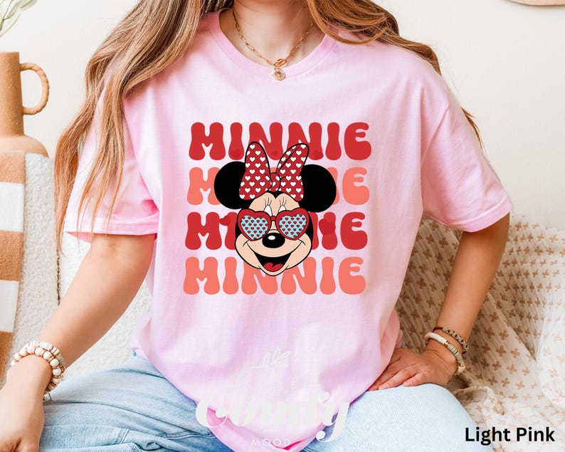 Puede incluir: Una camiseta rosa claro con un gr&aacute;fico retro de Minnie Mouse con gafas de sol en forma de coraz&oacute;n y un lazo rojo. El texto "MINNIE" se repite en un patr&oacute;n apilado.