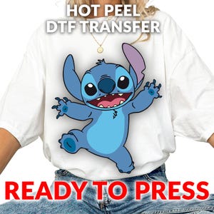 May include: White t-shirt featuring a cartoon image of Stitch, a blue alien character with large ears and a wide grin. The text "HOT PEEL DTF TRANSFER" and "READY TO PRESS" are printed above and below the character.