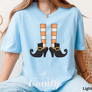 May include: Light blue t-shirt featuring a graphic of witch legs with orange and white striped stockings and black shoes with gold buckles. The text "Comfy Mood" is at the bottom of the shirt. The shirt is labeled "Light Blue".