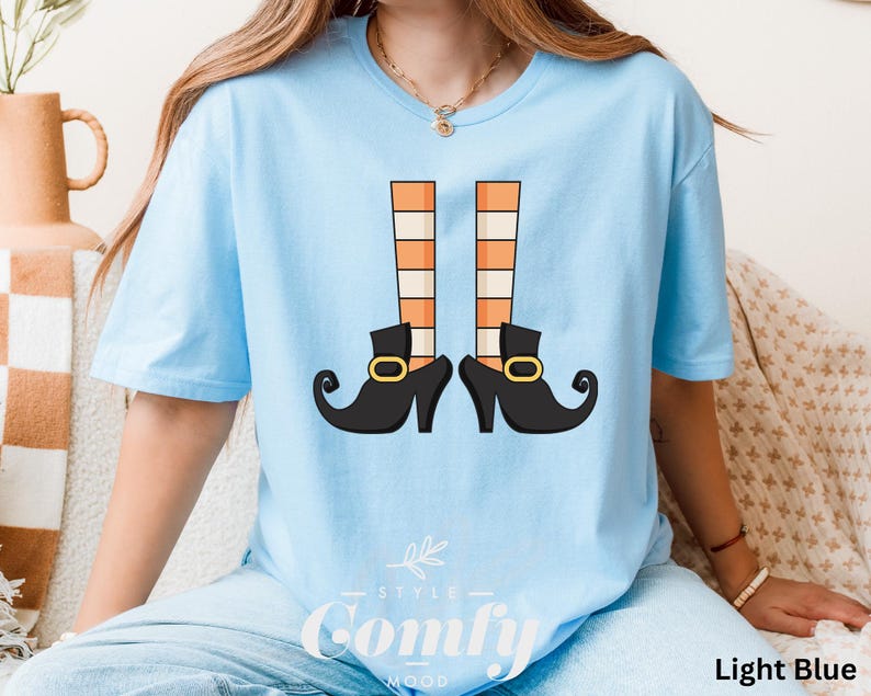 May include: Light blue t-shirt featuring a graphic of witch legs with orange and white striped stockings and black shoes with gold buckles. The text "Comfy Mood" is at the bottom of the shirt. The shirt is labeled "Light Blue".