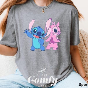May include: Sport grey t-shirt featuring a cartoon graphic of Stitch and Angel. Stitch is blue with large ears, and Angel is pink. The shirt has the words "STYLE" and "Comfy MOOD" printed on it.