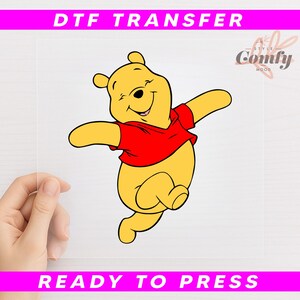 May include: A DTF transfer featuring a cartoon illustration of Winnie the Pooh in a red shirt, with the text "DTF TRANSFER" and "READY TO PRESS" in pink. The Comfy Mood logo is in the upper right corner.