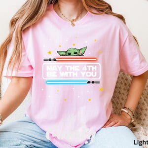 K&ouml;nnte beinhalten: Ein hellrosa T-Shirt mit einer gr&uuml;nen Alienfigur, die hinter einem roten und blauen Lichtschwert hervorlugt. Der Text "May the 4th Be With You" ist auf dem Shirt aufgedruckt.