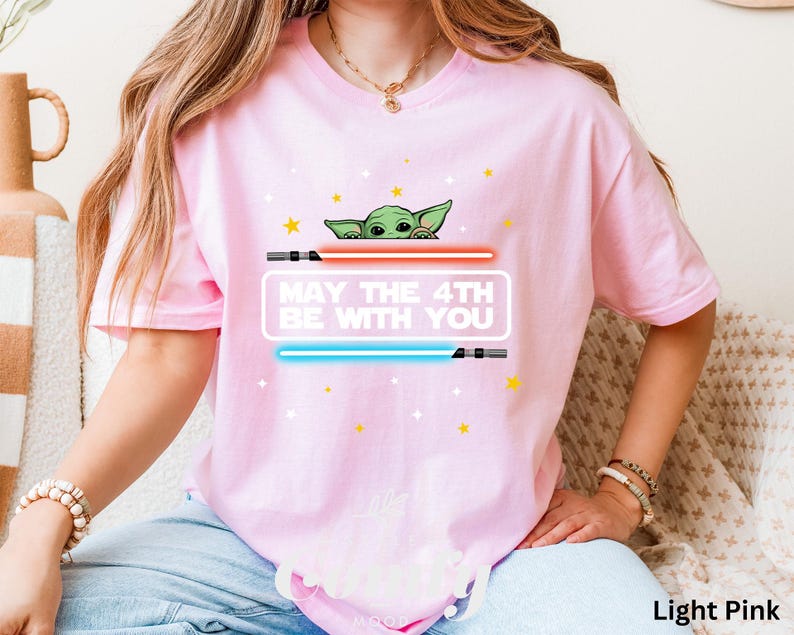 K&ouml;nnte beinhalten: Ein hellrosa T-Shirt mit einer gr&uuml;nen Alienfigur, die hinter einem roten und blauen Lichtschwert hervorlugt. Der Text "May the 4th Be With You" ist auf dem Shirt aufgedruckt.