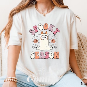 May include: Cream-colored t-shirt with the words "Spooky Season" in pink, orange, and white. A cartoon ghost with large eyes and a pumpkin is featured in the center. The shirt is a relaxed fit.