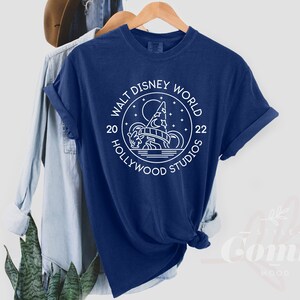 Puede incluir: Una camiseta azul marino con un dise&ntilde;o gr&aacute;fico blanco del logotipo de Walt Disney World Hollywood Studios. El logotipo presenta una imagen estilizada del Hollywood Tower Hotel con una luna y estrellas en el fondo. El texto "Walt Disney World" est&aacute; escrito en un c&iacute;rculo alrededor de la imagen, con "20" y "22" a cada lado del c&iacute;rculo. El texto "Hollywood Studios" est&aacute; escrito debajo del c&iacute;rculo.